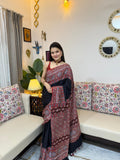 A woman wearing a deep black modal silk Ajrakh saree with intricate red and indigo block prints on the pallu and border, accessorized with a red choker necklace.