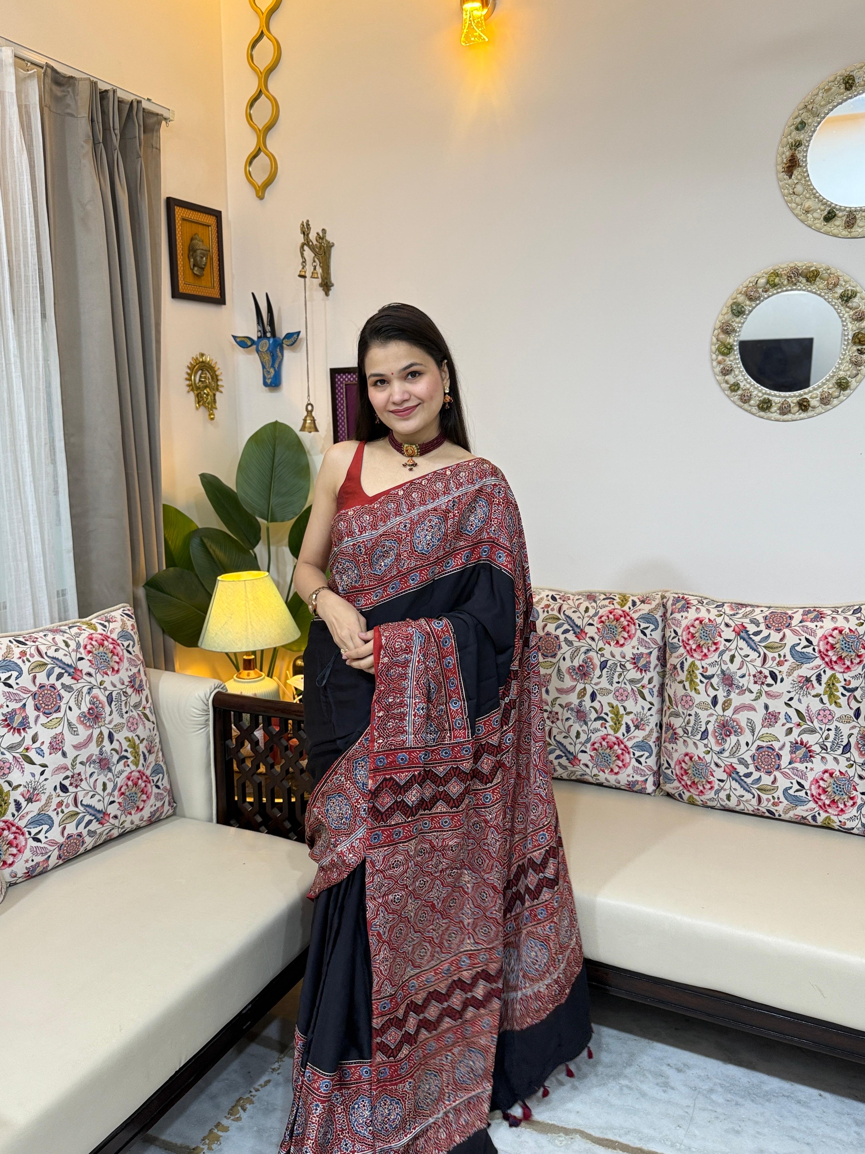 A woman wearing a deep black modal silk Ajrakh saree with intricate red and indigo block prints on the pallu and border, accessorized with a red choker necklace.