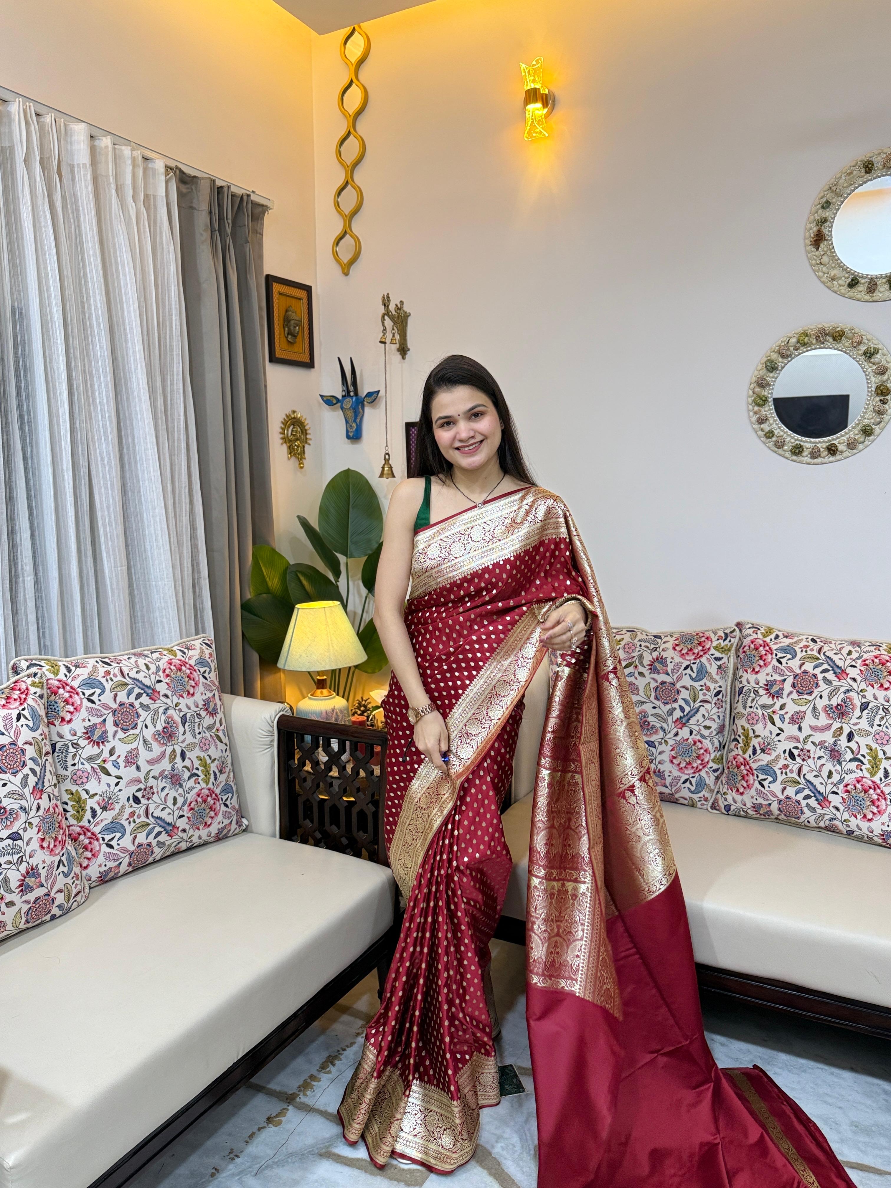 A woman is gracefully modeling a deep maroon Katan silk Banarasi saree from Rivaz, featuring intricate gold zari buttis throughout the body and a traditional woven border.