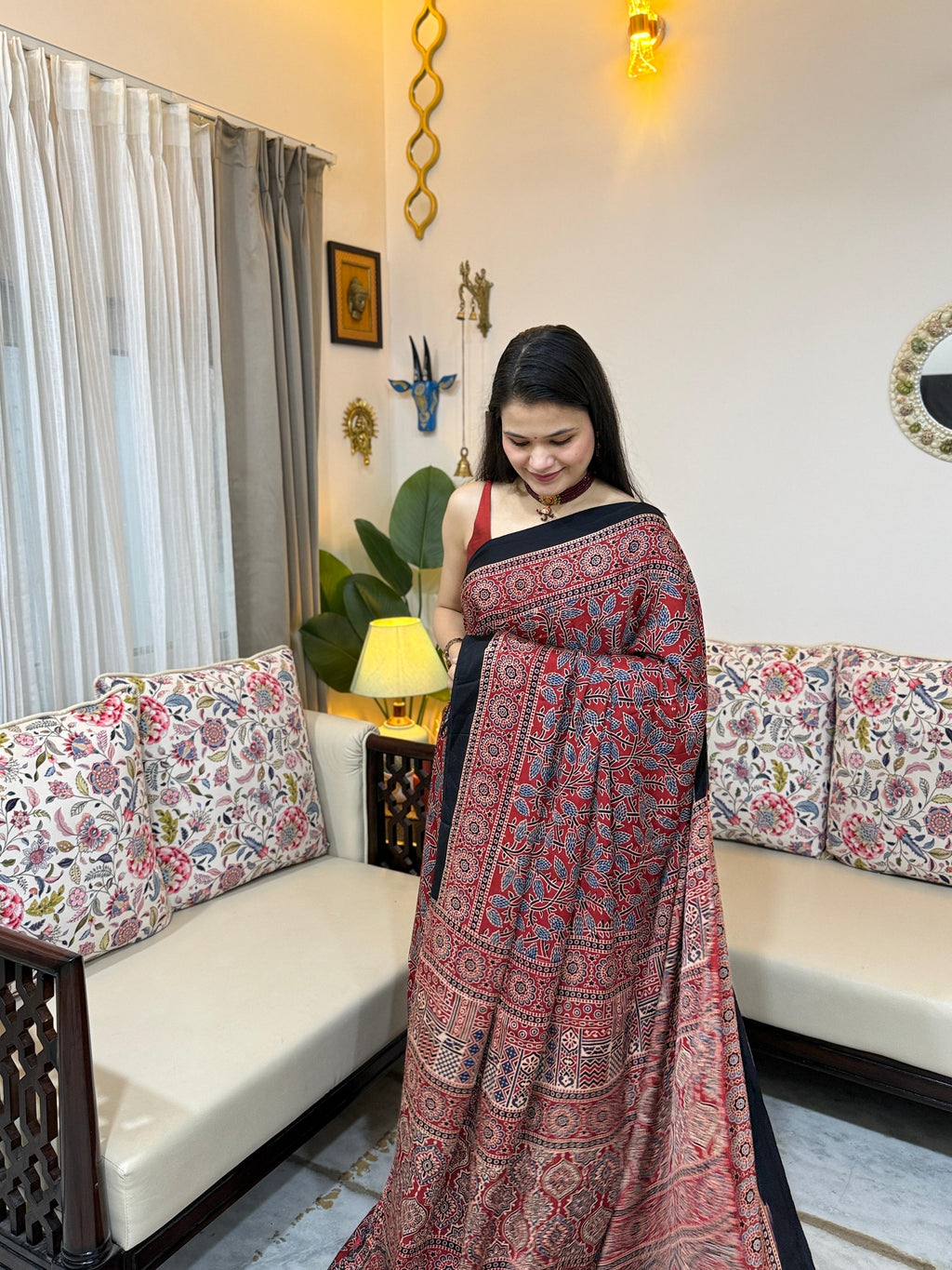 A woman draping a crimson red modal silk saree featuring all-over traditional Ajrakh block prints and a matching printed pallu, styled with a red choker necklace.