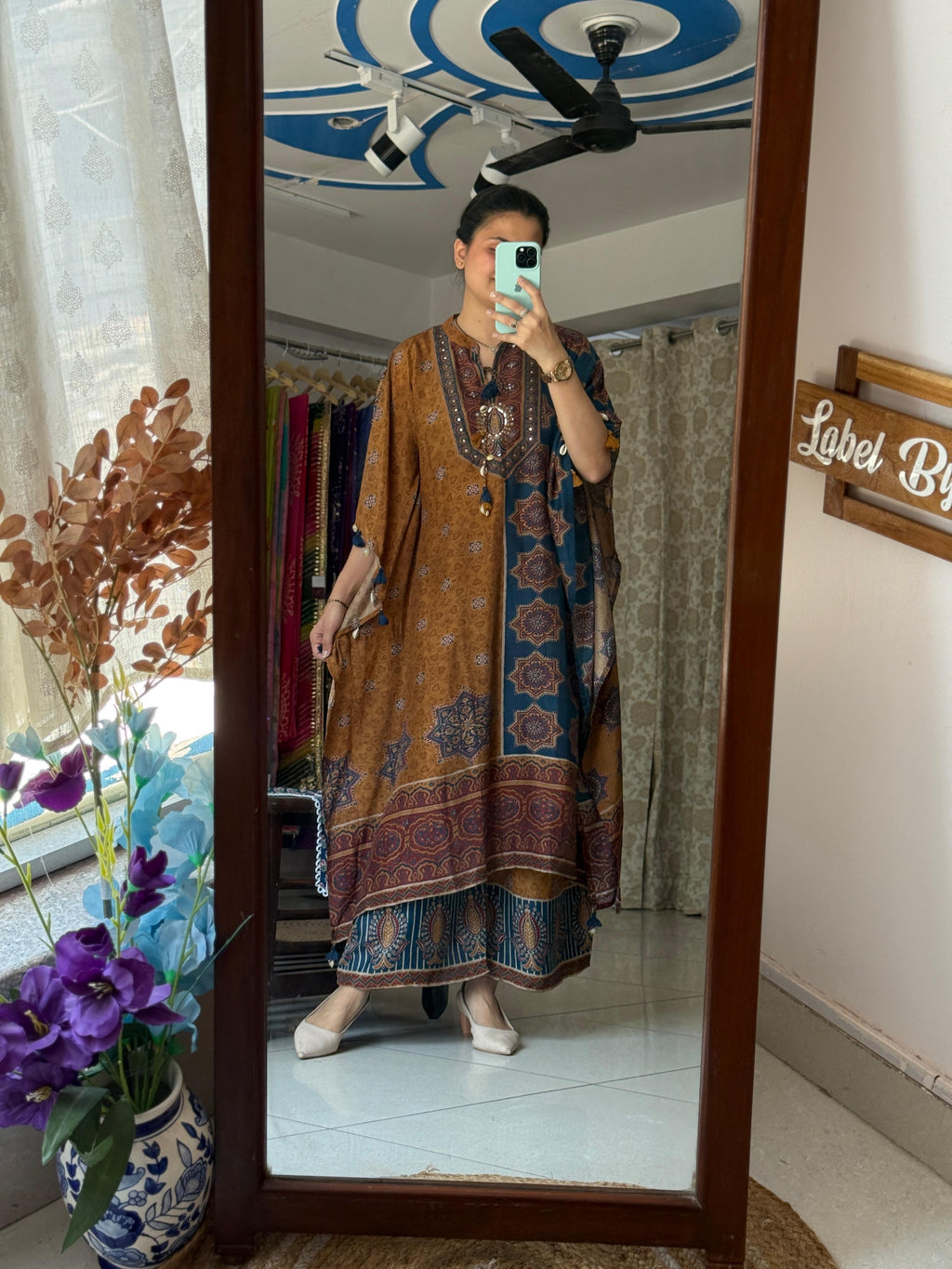 A woman taking a mirror selfie wearing a mustard brown and teal blue printed pure muslin kaftan co-ord set featuring a mirror work neckline, cowrie shell tassels, and matching wide-leg pants.